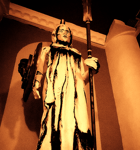 Statue imposante d'un guerrier ou gardien en pierre, vêtu d'une toge, tenant lance et bouclier, dans une arche sombre et chaude.
