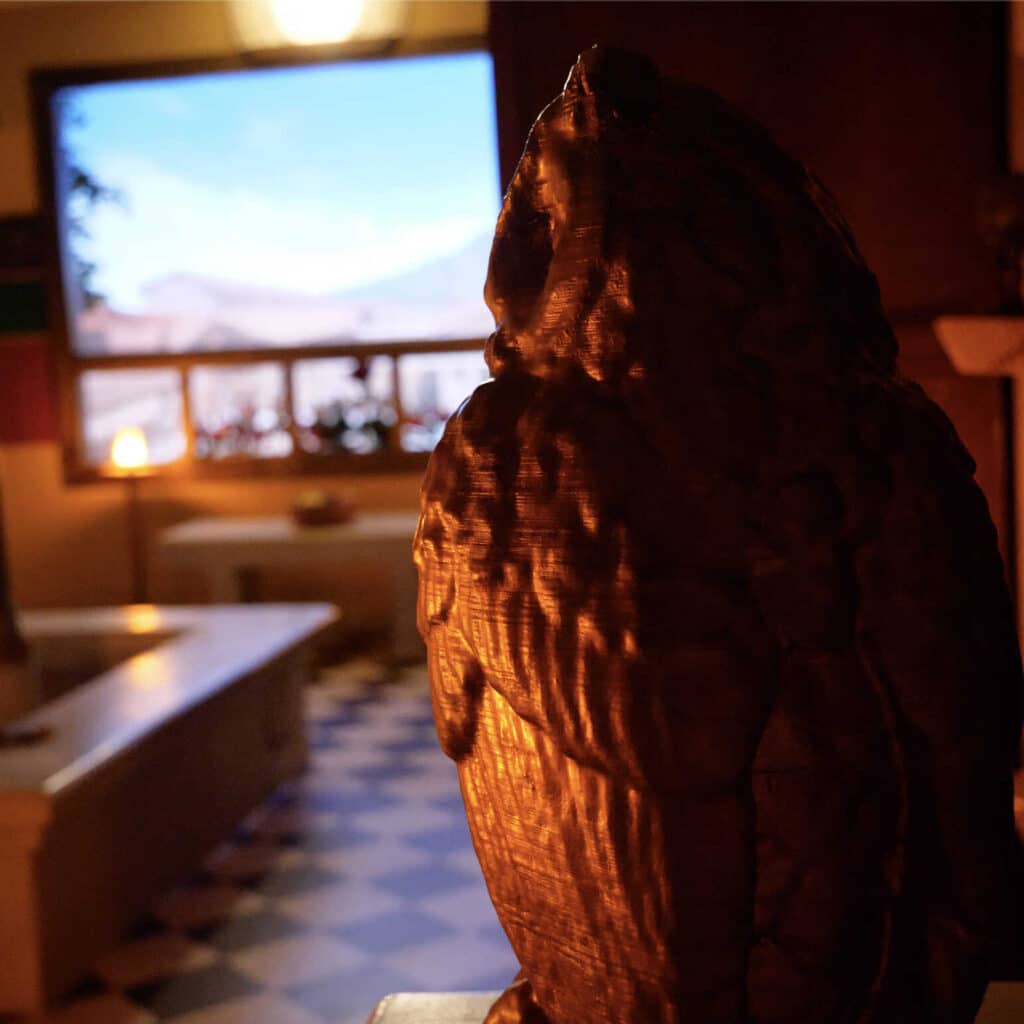 Statue d'hibou en bronze foncé, texture visible, éclairée. En arrière-plan flou, une fenêtre encadre un ciel bleu et des montagnes, avec un intérieur chaleureux.